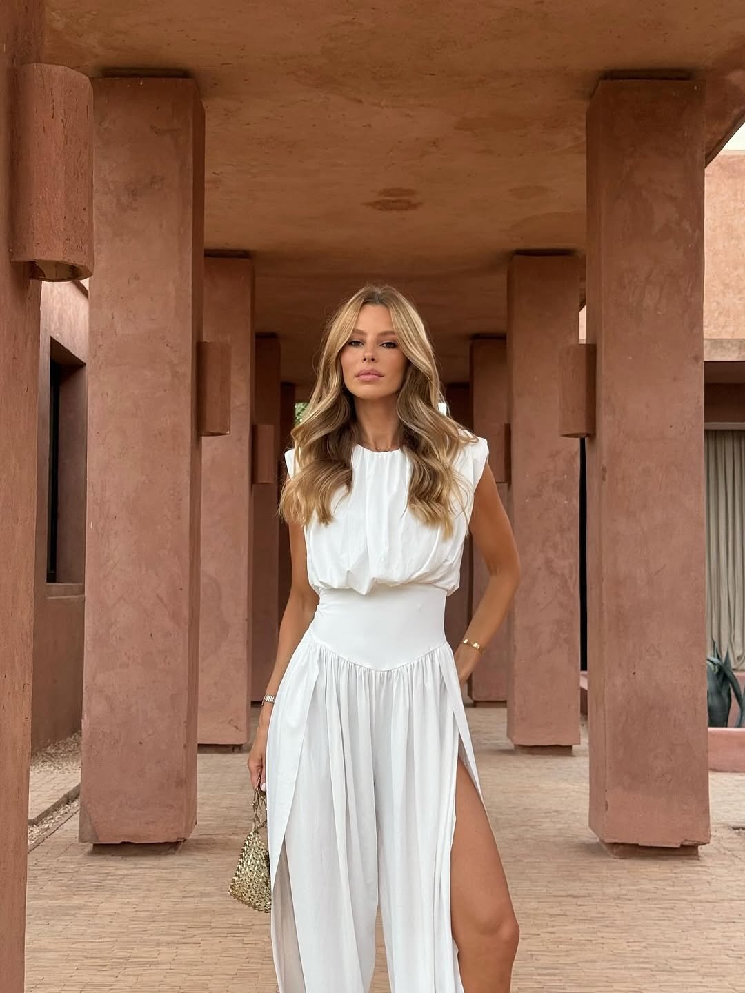 Mulher com macac&atilde;o branco fluido, fenda elegante, em corredor de arquitetura rosada, estilo sofisticado e leve, ideal como look para