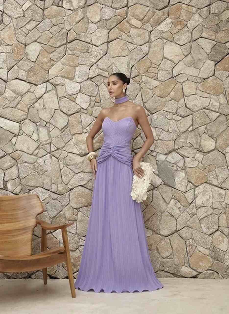 Pessoa em frente a parede de pedra usando vestido para casamento no ver&atilde;o lavanda, tomara que caia, longo e plissado, com clutch floral, acess&oacute;rios el