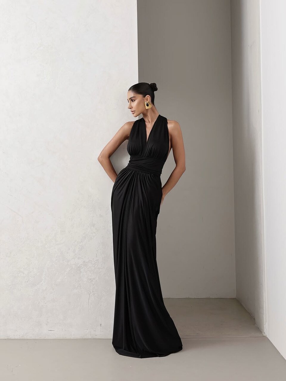 Mulher em vestido preto longo, destacando tecidos para vestido de festa sofisticados, ambiente minimalista claro, acess&oacute;rios dourados modernos.